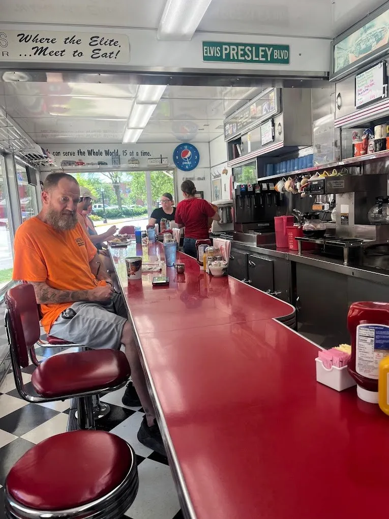The long red counter and swivel stools under the “Where the Elite… Meet to Eat!” sign and an Elvis Presley Blvd street sign