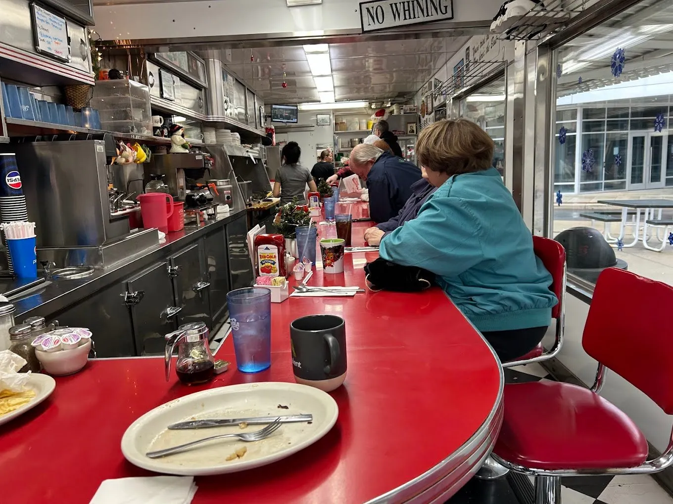 Regulars at the curved red counter, looking toward the grill, under a “No Whining” sign